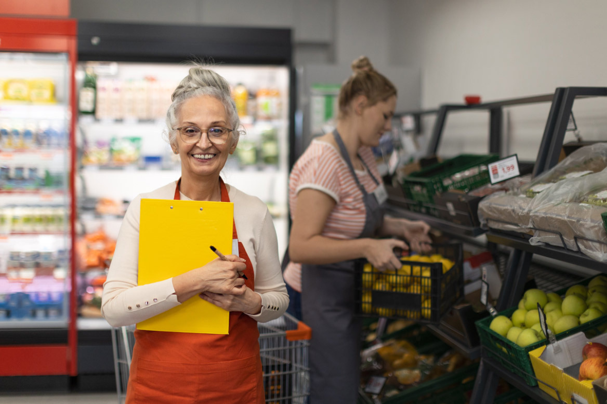 Two women working in a social supermarket; one holding a clipboard and smiling, while the other arranges produce on shelves.